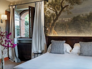 Un dormitorio con una cama grande, una ventana con cortinas, una mesita con una taza, y un cuadro en la pared.