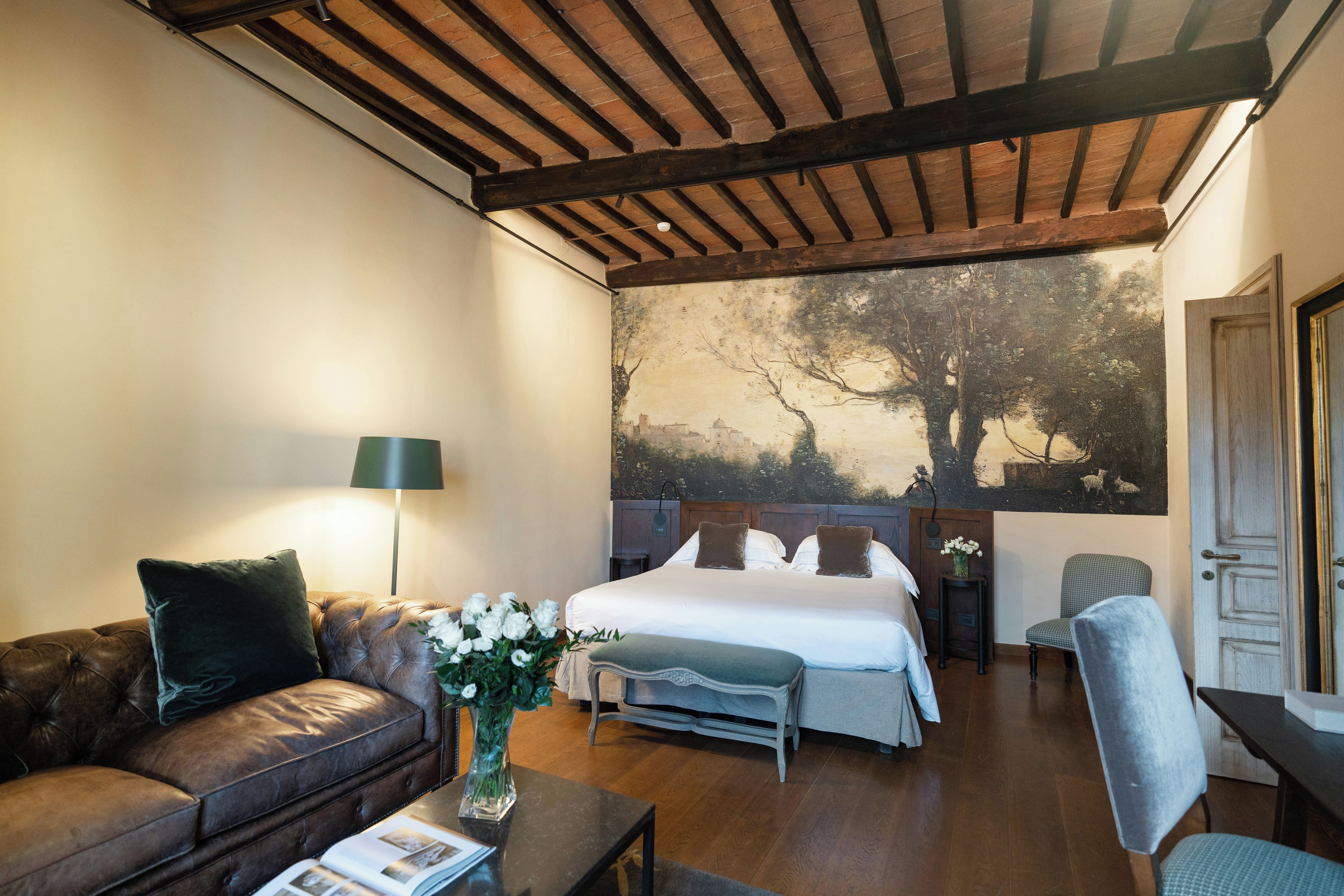 Una camera da letto con un letto grande, un'area salotto con divano e poltrona, un comodino con lampada e un muro decorato con un murale raffigurante un albero.