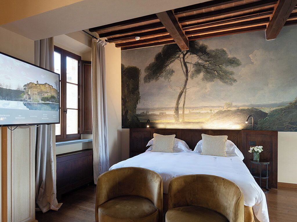 A hotel room with a bed, two chairs, a painting on the wall, a TV on the left, and a window with curtains.