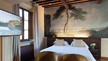 A hotel room with a bed, two chairs, a painting on the wall, a TV on the left, and a window with curtains.