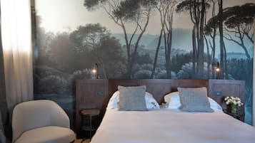 Una camera da letto con un letto grande, due cuscini, una sedia, un comodino con un vaso di fiori e un murale raffigurante alberi sulla parete.
