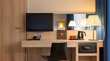 A hotel room with a wooden desk, a flat-screen TV, a lamp, a coffee maker, and a minibar.