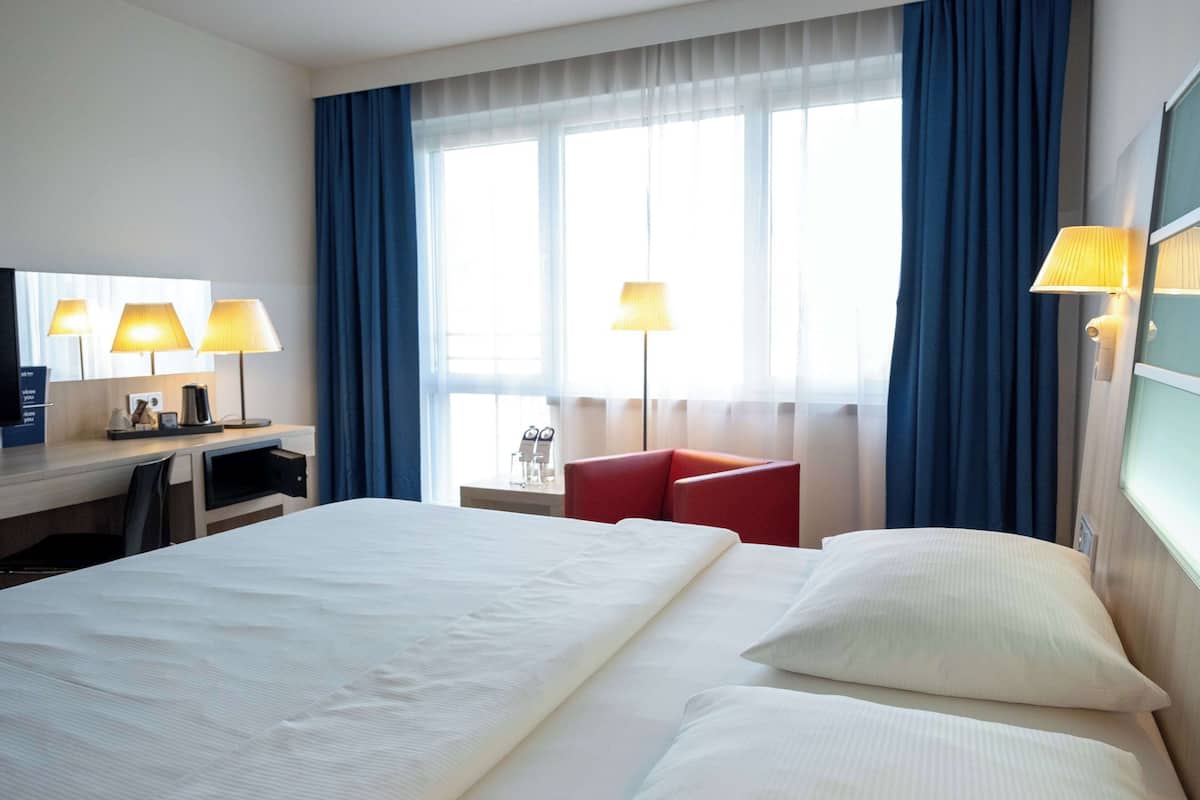 A hotel room with a large bed, a desk with a chair, two lamps, and a red armchair.