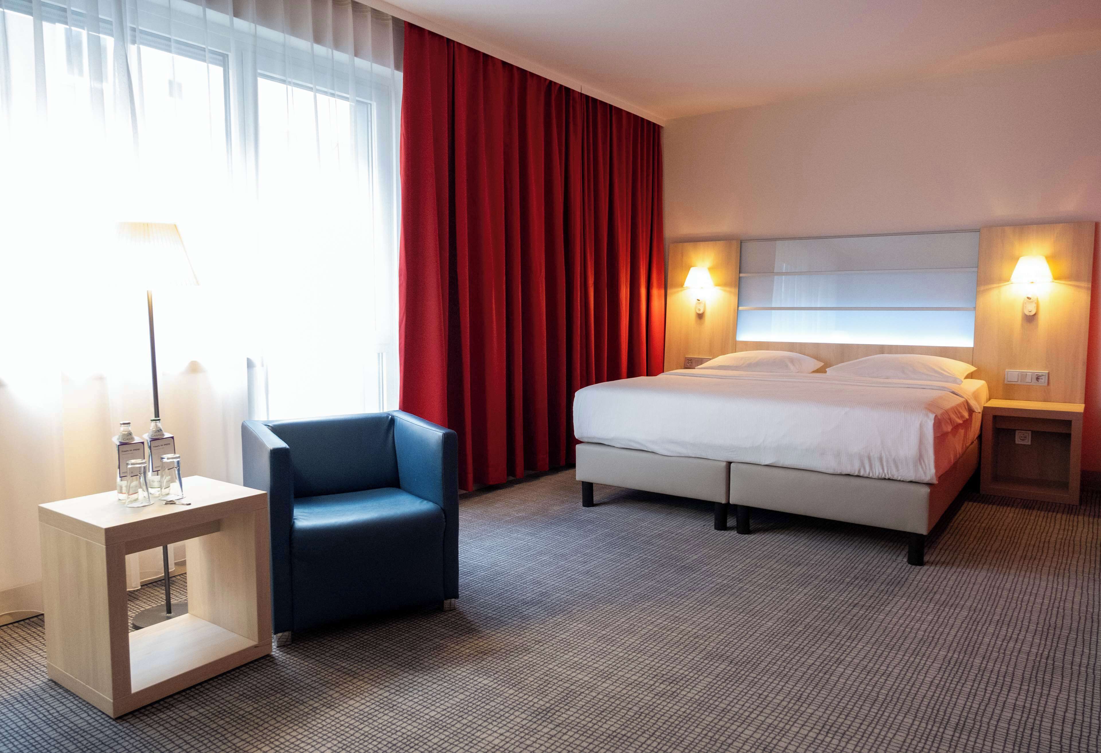 Ein Hotelzimmer mit Bett, Stuhl, Nachttisch mit Lampe und einem Fenster mit Vorhängen.