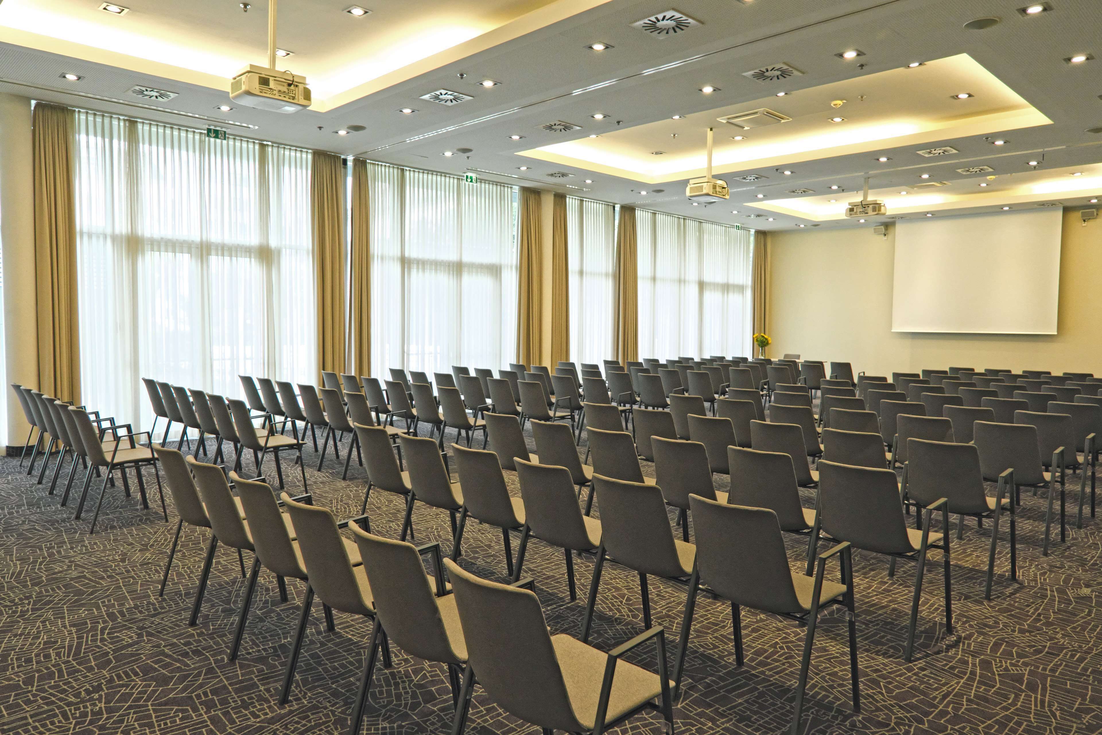 Un salón de conferencias con filas de sillas, una pantalla de proyector y piso alfombrado.