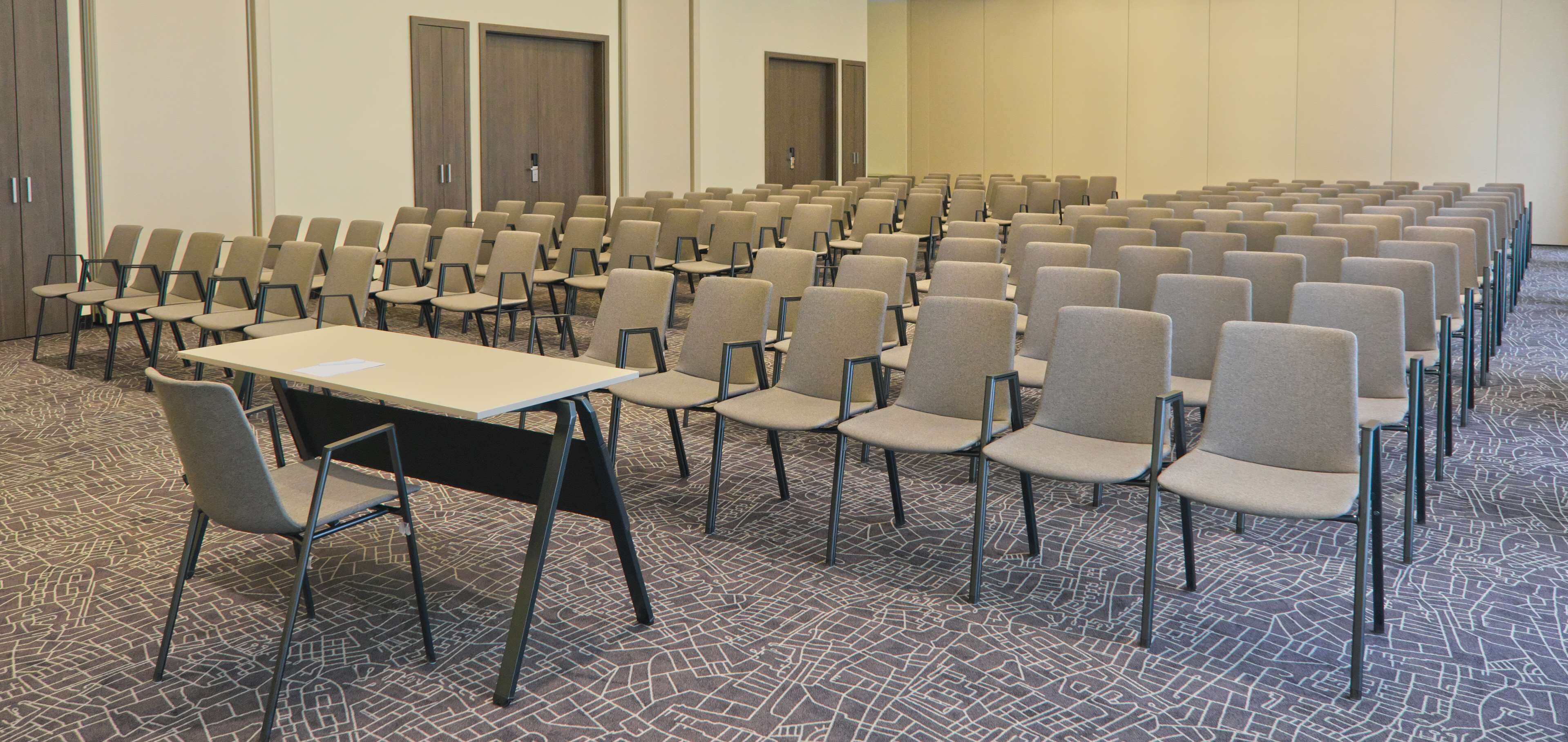 Une salle de conférence avec des rangées de chaises, une table et des portes au fond.