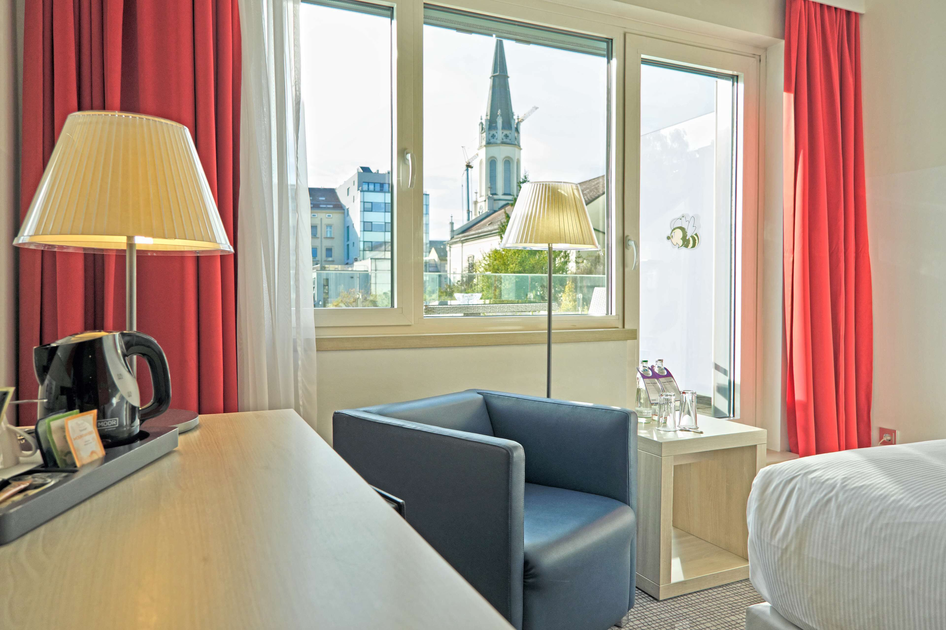 Ein Hotelzimmer mit einem Bett, einem Schreibtisch mit Lampe, einem Sessel und Blick auf eine Kirche durch das Fenster.