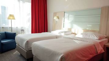 A hotel room with two beds, a blue armchair, a desk, and a window with curtains.