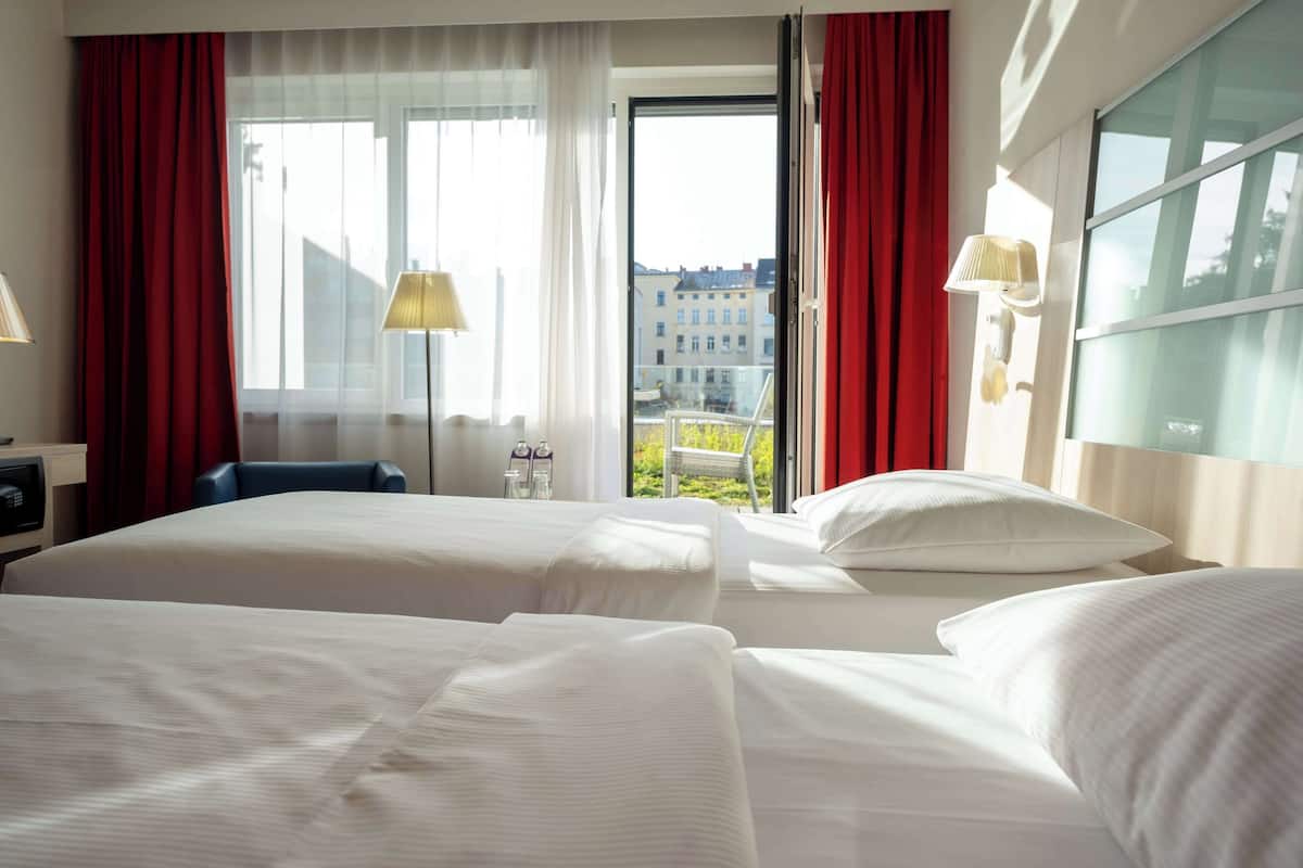A hotel room with two beds, a desk, a chair, a lamp, and a view of buildings through the window.