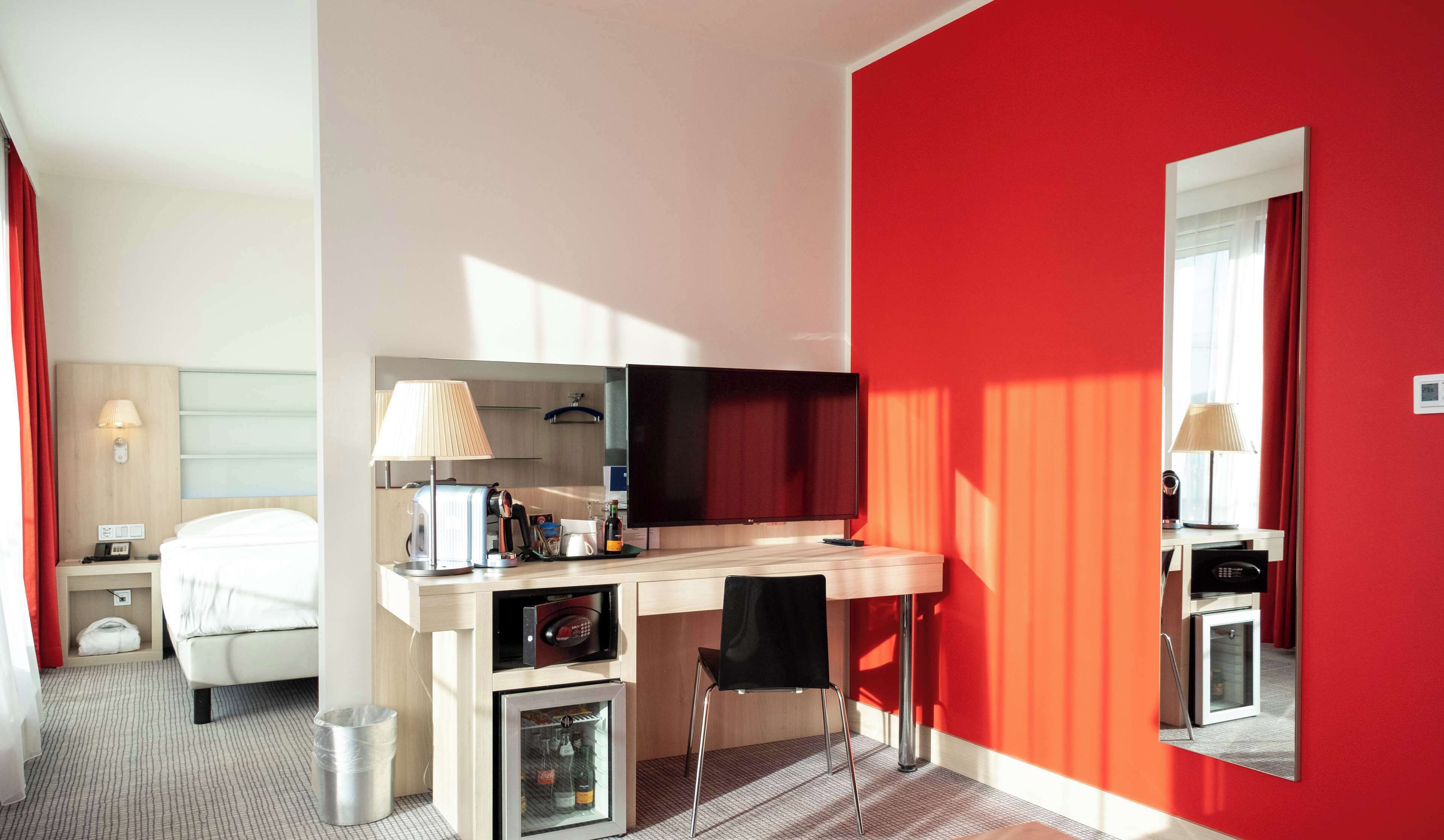 A hotel room with a bed, a desk with a lamp, a small refrigerator, and a mirror on the wall.