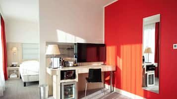 A hotel room with a bed, a desk with a lamp, a small refrigerator, and a mirror on the wall.