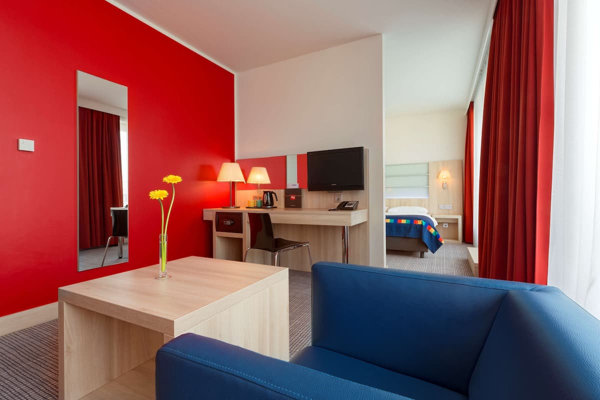 A hotel room with a red wall, a desk with a chair, a TV, a bed with a blue bedspread, and a blue sofa.