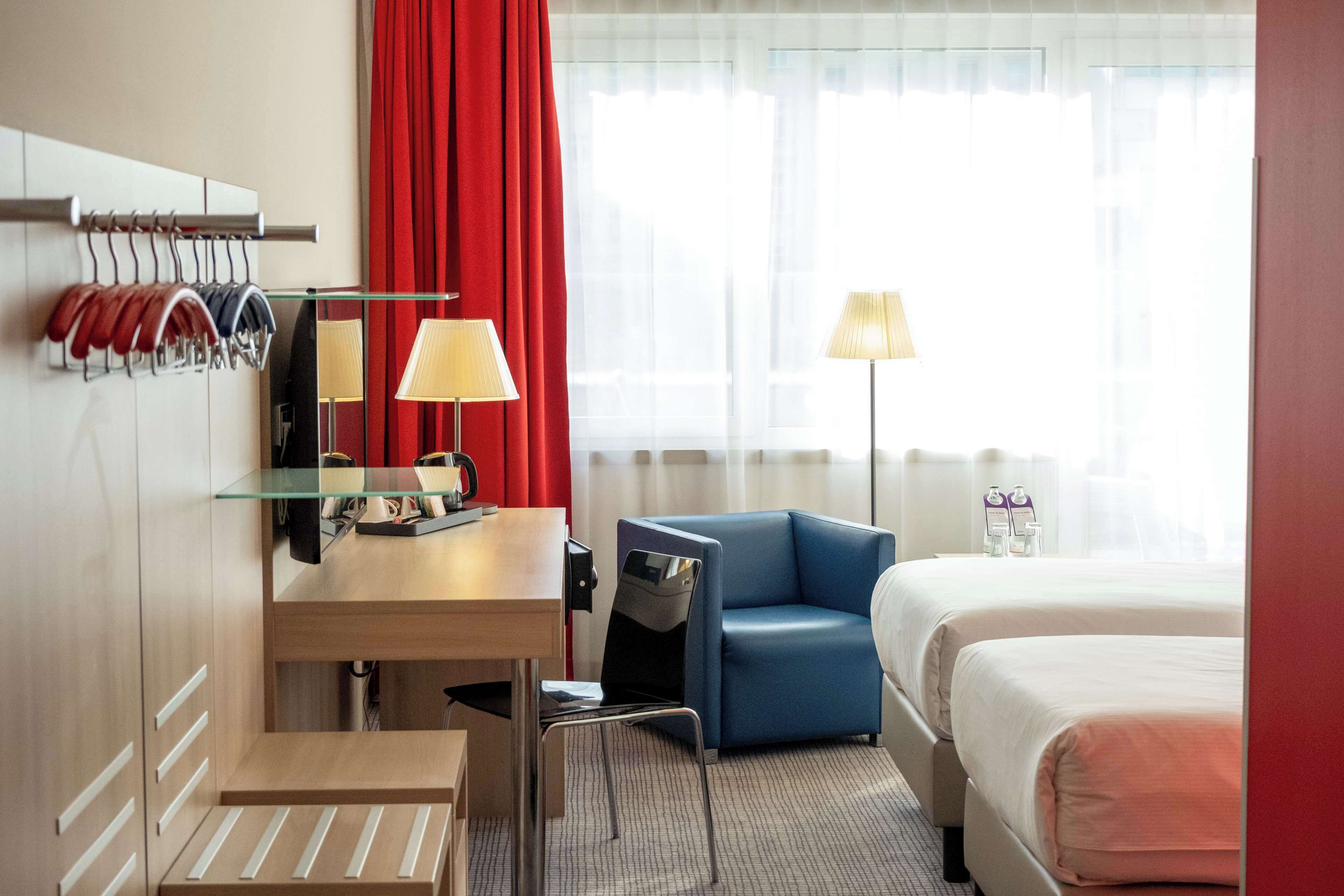 A hotel room with a bed, a desk with a chair, a blue armchair, a lamp, and a coat rack with hanging items.