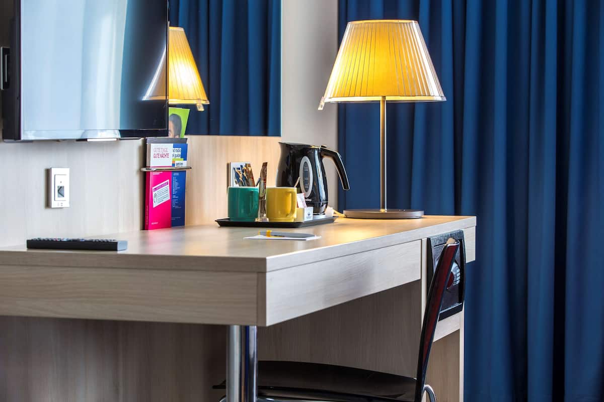 A hotel room with a wooden desk, two lamps, a kettle, a mug, and a remote control.