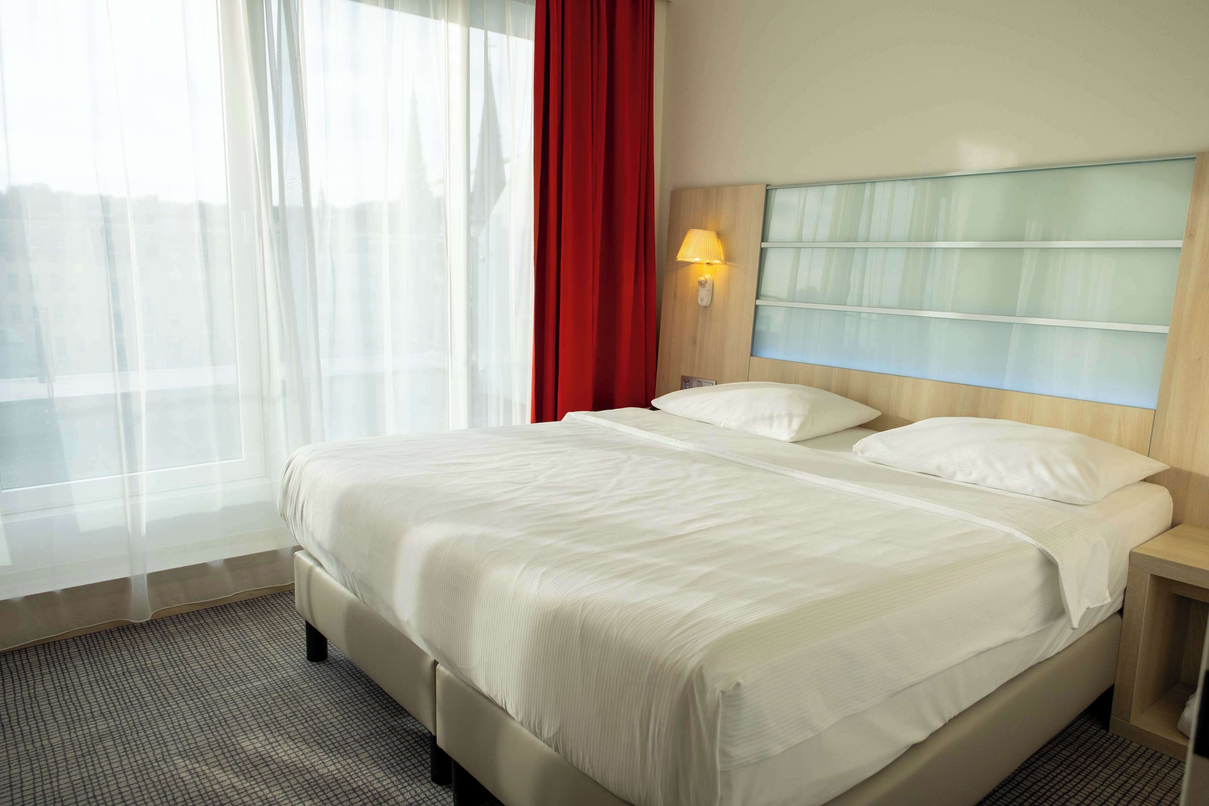 Una habitación de hotel con una cama, una mesita de noche, una lámpara, una ventana con cortinas translúcidas y una cortina roja.