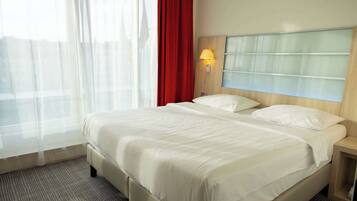 Una habitación de hotel con una cama, una mesita de noche, una lámpara, una ventana con cortinas translúcidas y una cortina roja.