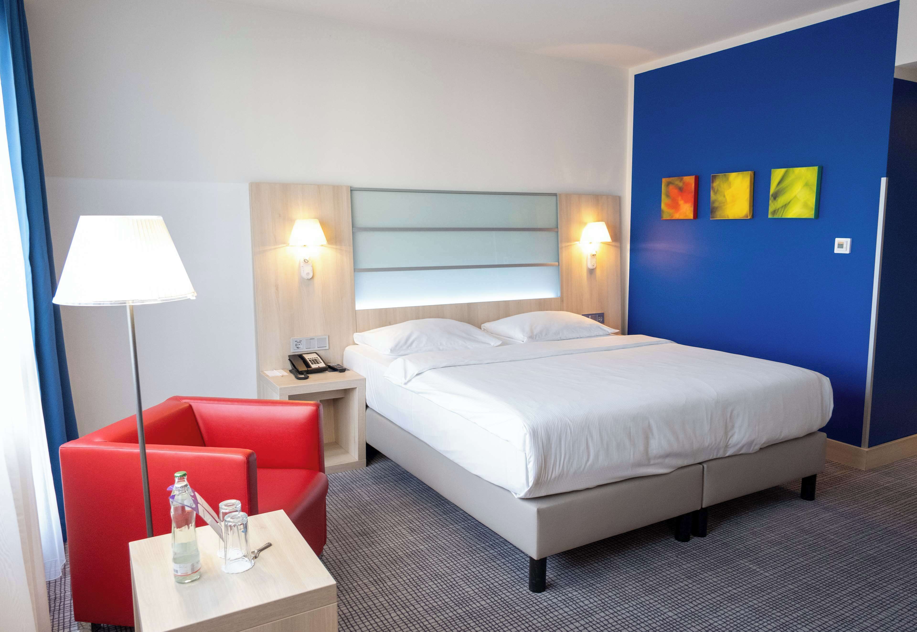 A hotel room with a bed, a red armchair, a nightstand with a lamp, and a wall with three paintings.