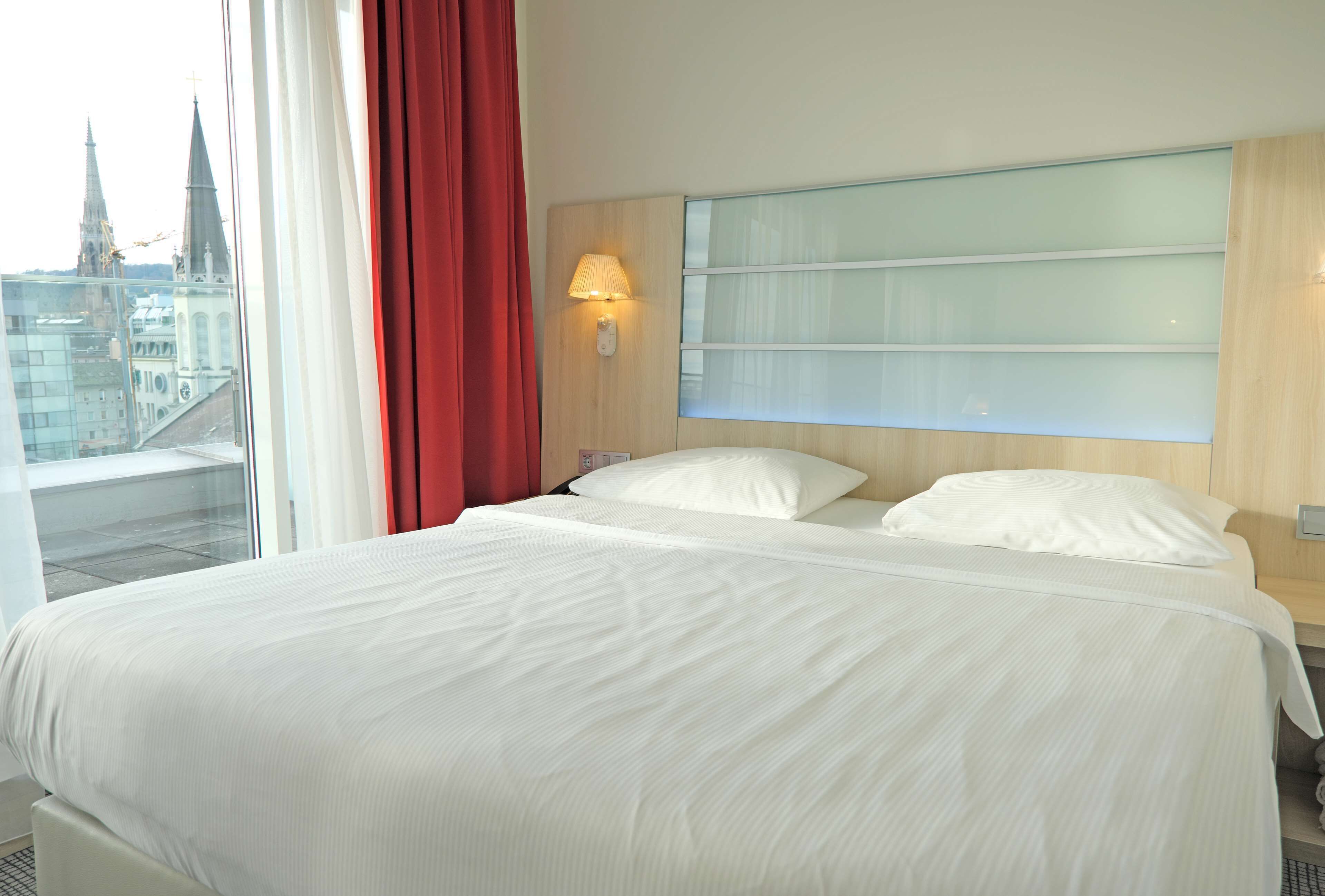 A hotel room with a large bed, a nightstand, a wall-mounted lamp, and a view of city buildings.