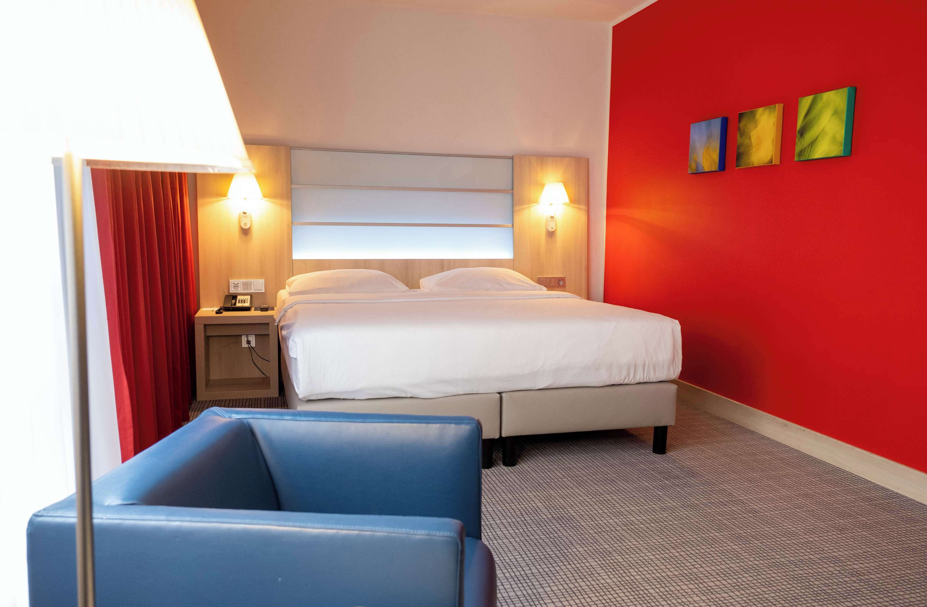 A hotel room with a bed, a nightstand, a blue chair, and a red accent wall.