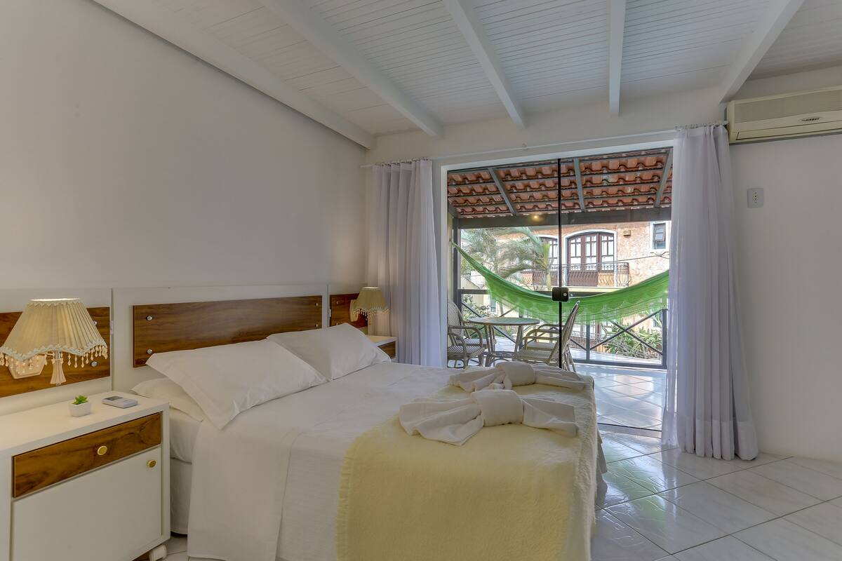Habitación de hotel con cama, mesita de noche, lámpara y vistas a un patio con hamaca y sillas.