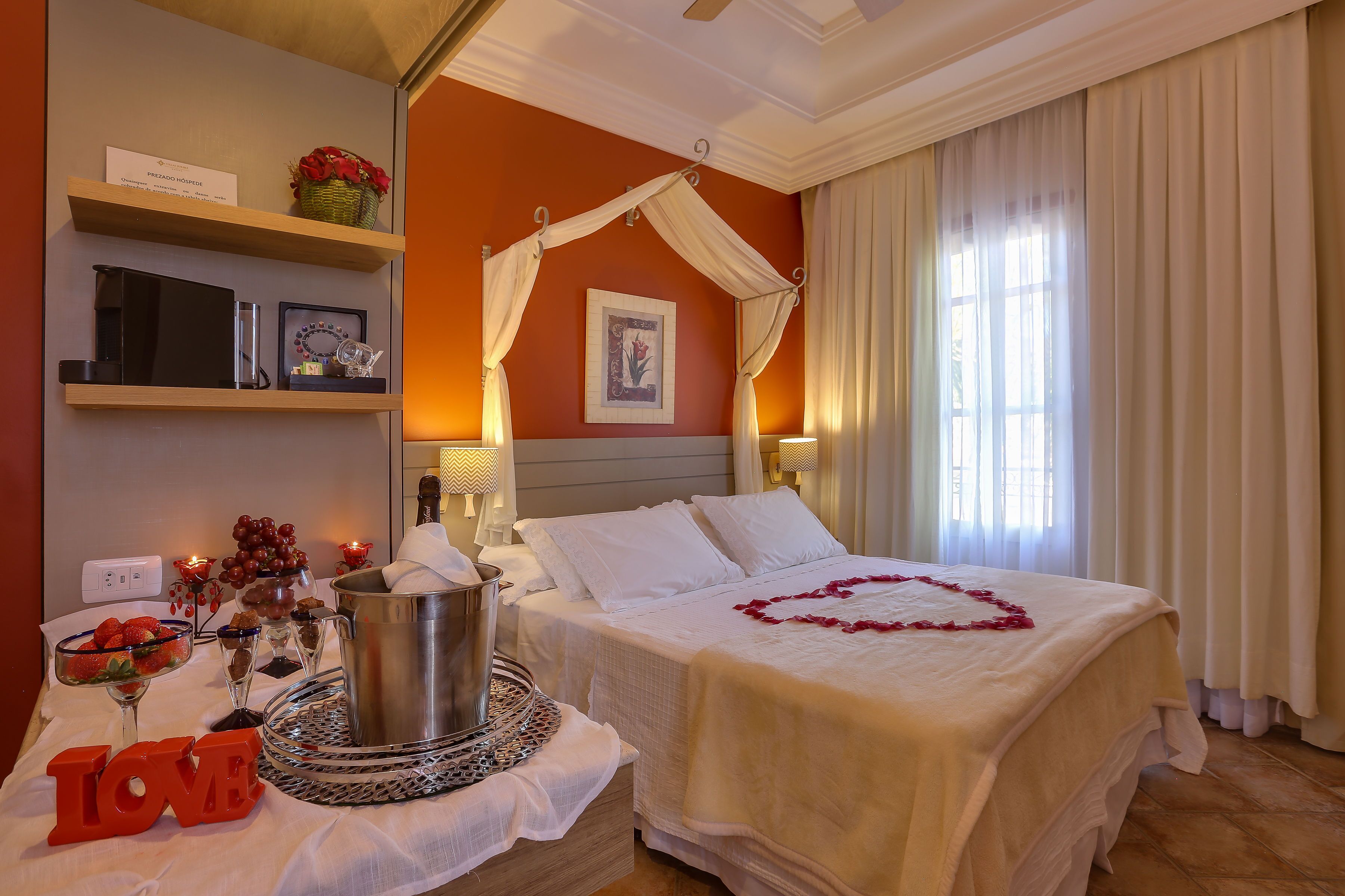 A hotel room with a bed, a bedside table with a 'LOVE' sign, a vase with flowers, a framed picture, and a shelf with a basket and a sign.