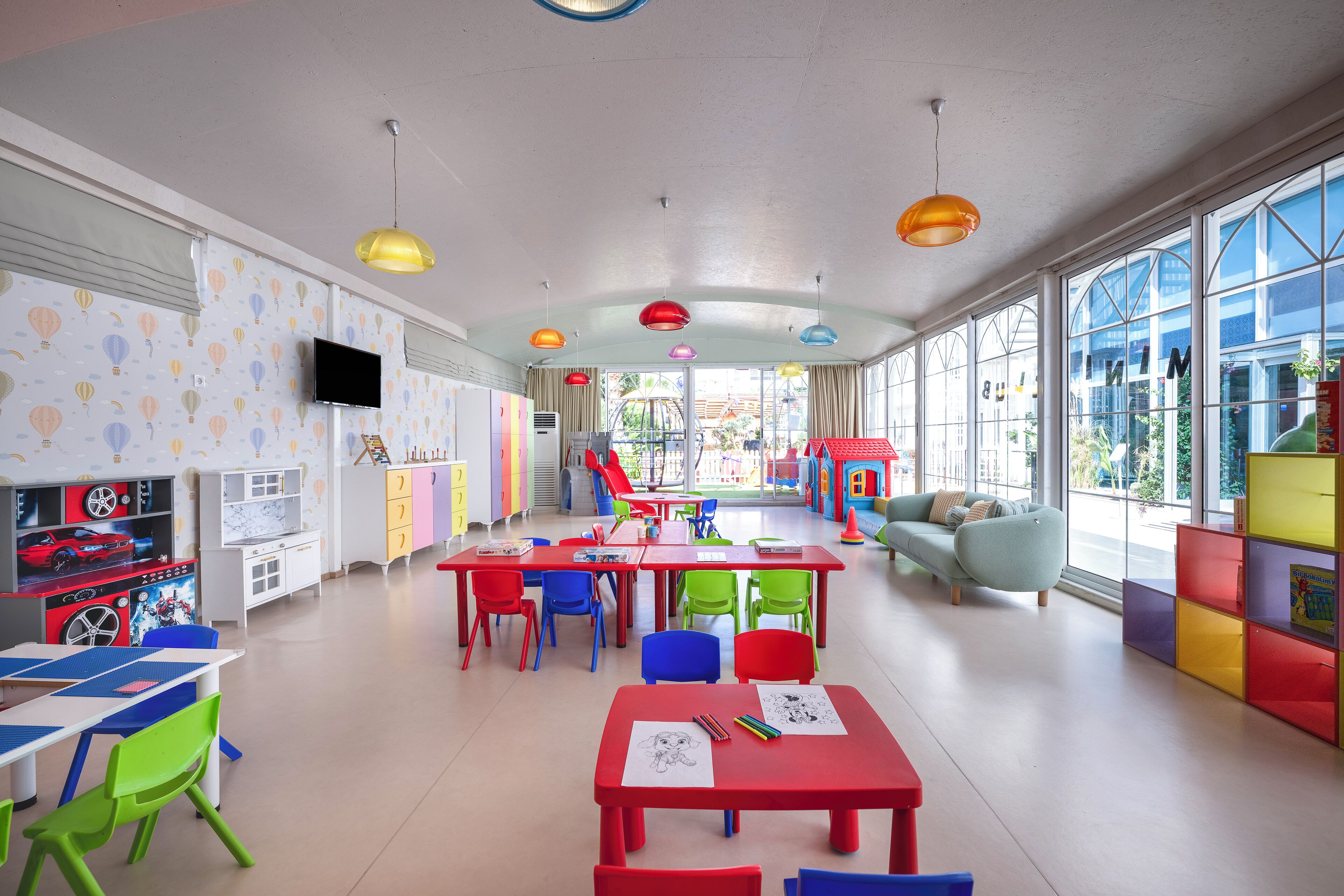 Une salle de jeux pour enfants aux couleurs vives, avec des tables, des chaises et des espaces dédiés aux jeux.