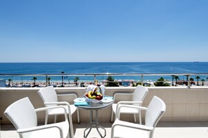 A balcony with white wicker chairs and a small round table with fruit and a bottle. The view includes a beach and the ocean.