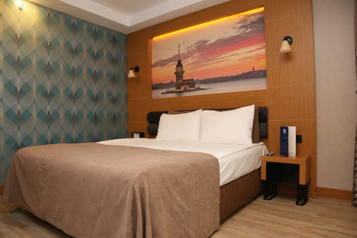 Ein Hotelzimmer mit einem Bett, einem Nachttisch, einer Wandleuchte, einem gemusterten Wandteppich und einem Sonnenuntergangsbild.