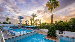 A swimming pool with lounge chairs and umbrellas, surrounded by palm trees and a sunset sky.