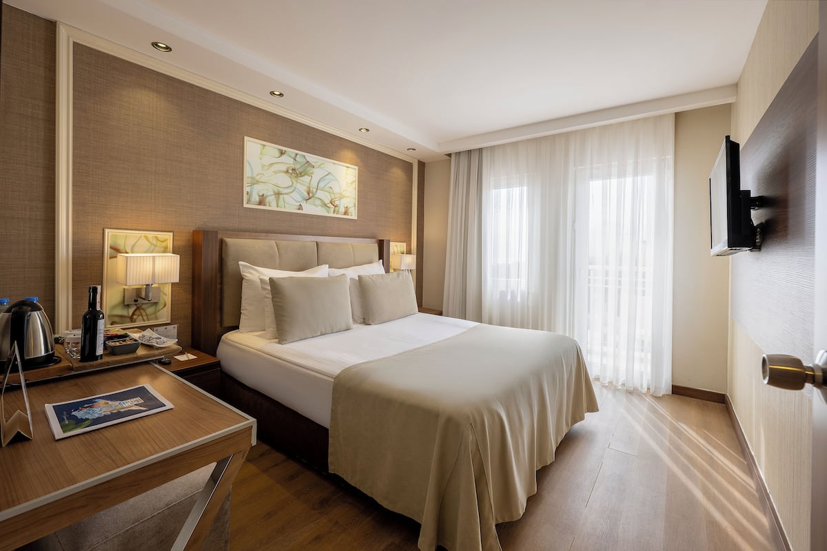 Ein Hotelzimmer mit einem großen Bett, einem Nachttisch mit Lampe, einem an der Wand befestigten Flachbildfernseher und einem Fenster mit durchsichtigen Vorhängen.