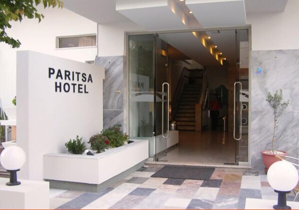 The entrance to Paritsa Hotel with a modern design, featuring a glass door, a staircase, and a reception area with a white wall and potted plants.