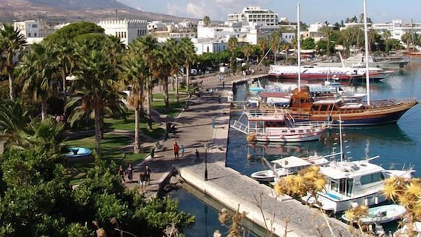 A marina with boats docked, a promenade lined with palm trees, and buildings in the background. - Paritsa Hotel (Kos)