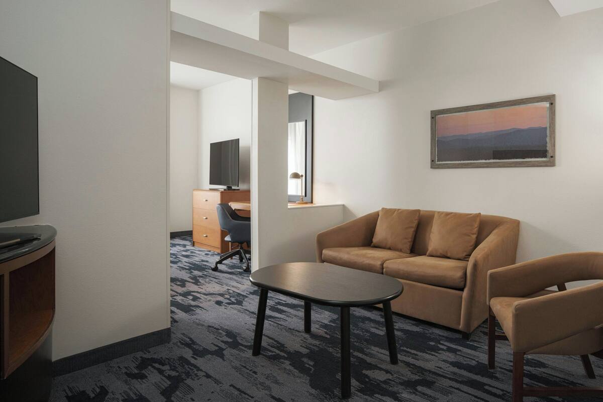 A hotel room with a sofa, armchair, and a small table. There is a desk with a chair and a television in the corner.