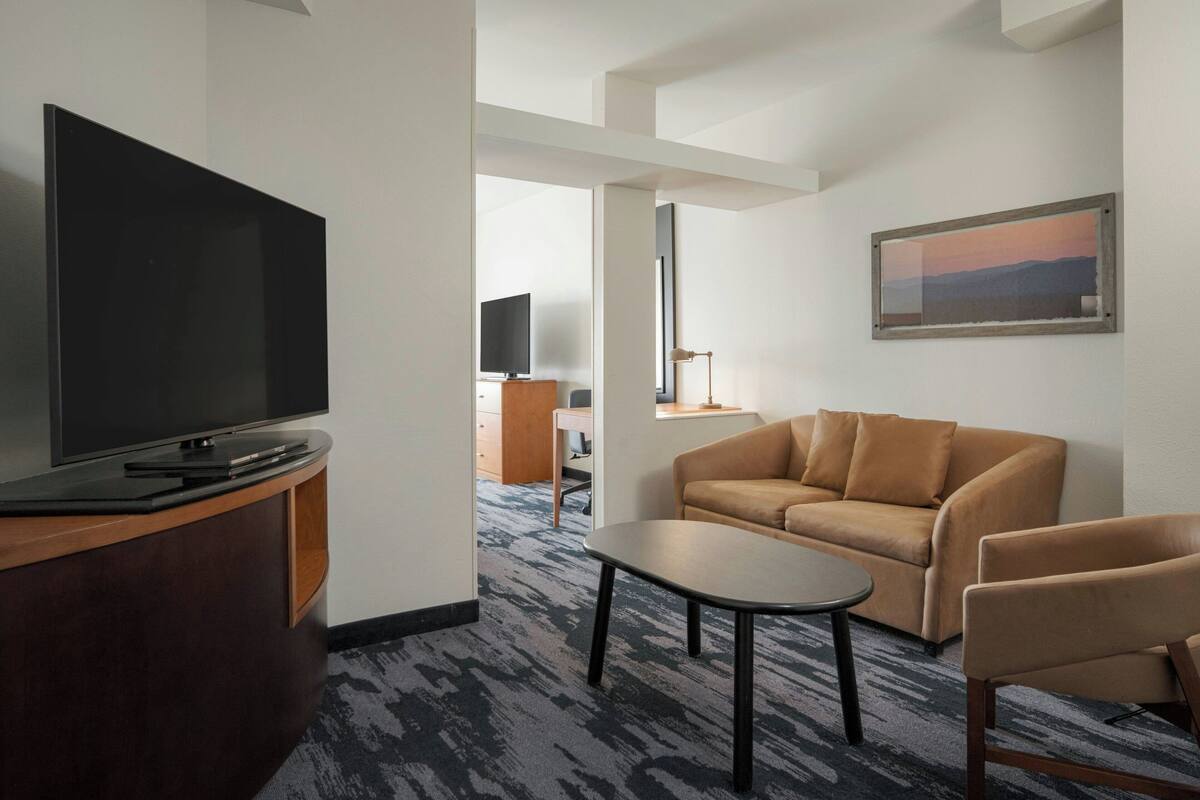 A hotel room with a flat-screen TV on a wooden stand, a brown sofa, a small table, and a framed picture on the wall.