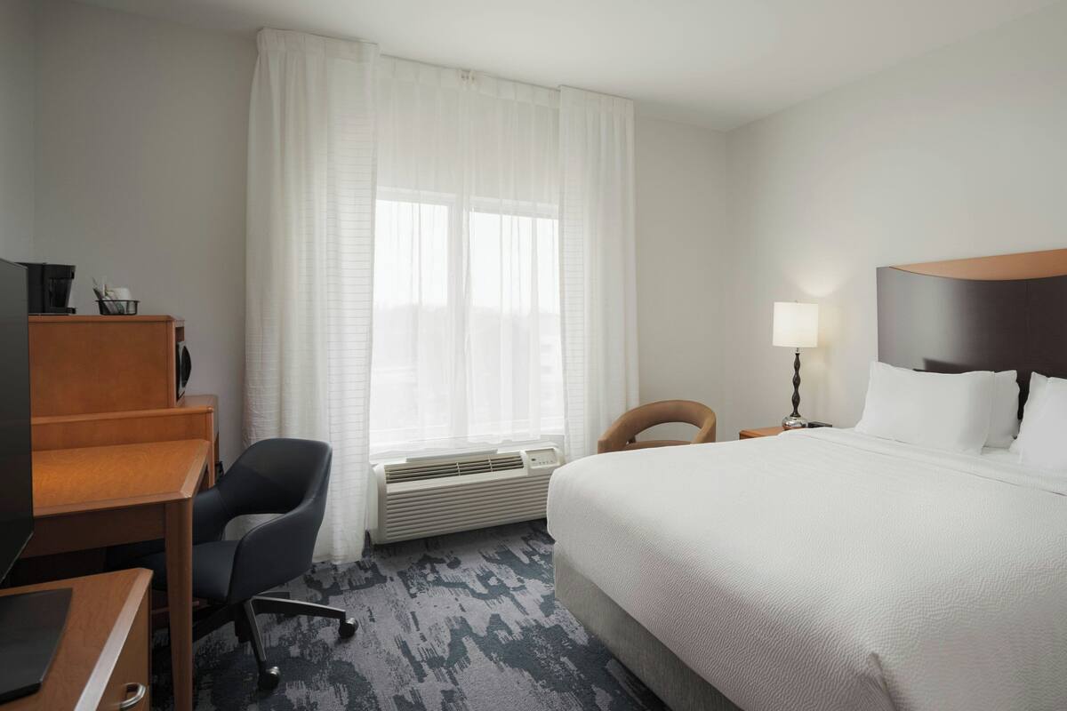 A hotel room with a bed, a desk, a chair, a lamp, and a window with curtains.
