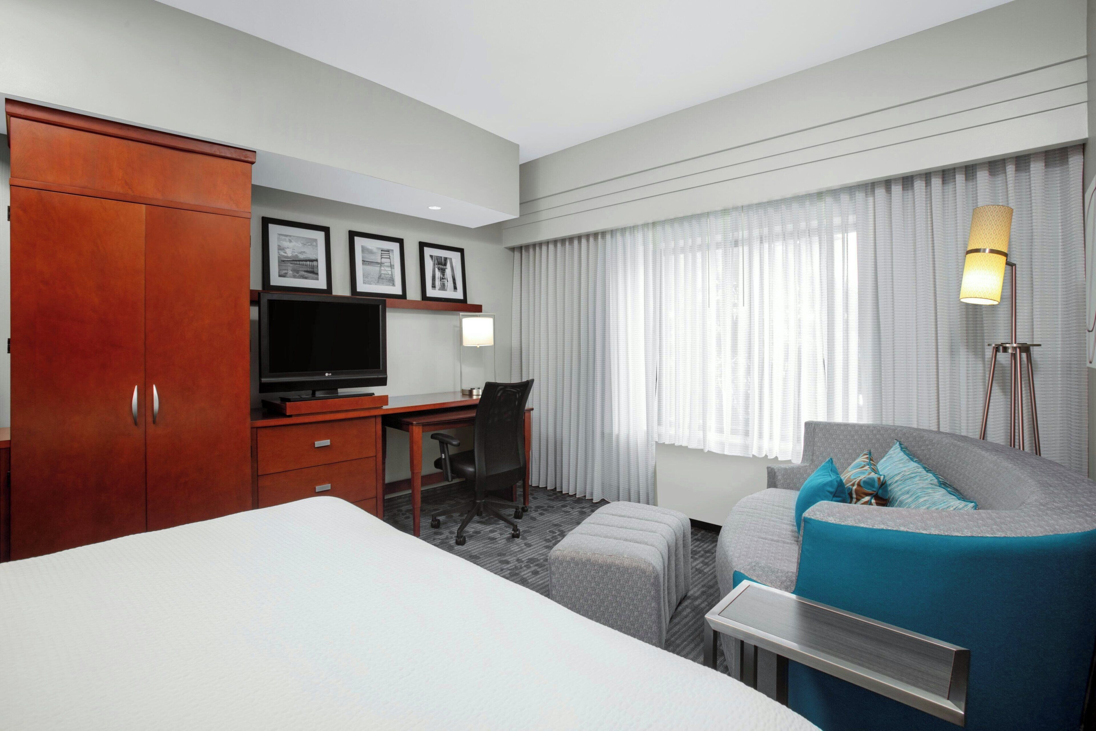 Quarto de hotel com cama, escrivaninha com cadeira, televisão, sofá e janela com cortinas.