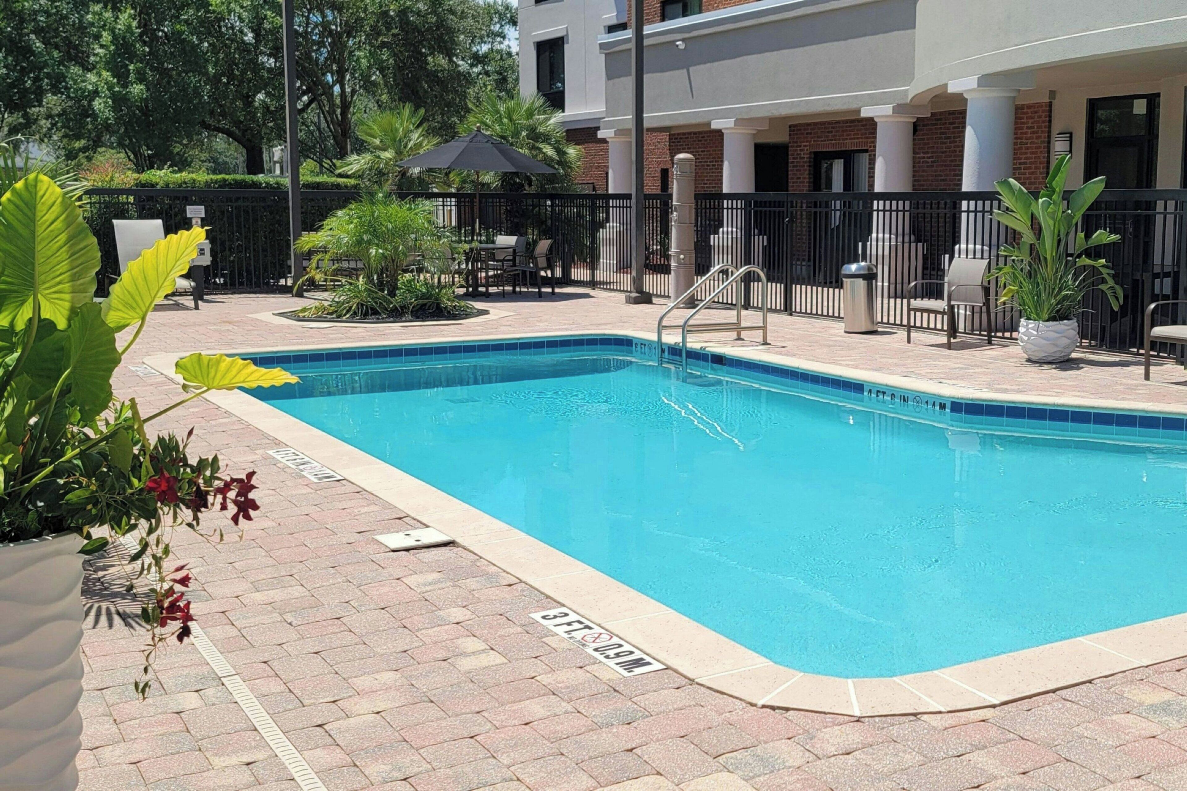 Piscina com borda azulejada, cercada por um pátio de tijolos e uma cerca de metal preto. Vasos com plantas e móveis de exterior à vista.