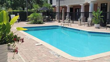 Piscina com borda azulejada, cercada por um pátio de tijolos e uma cerca de metal preto. Vasos com plantas e móveis de exterior à vista.
