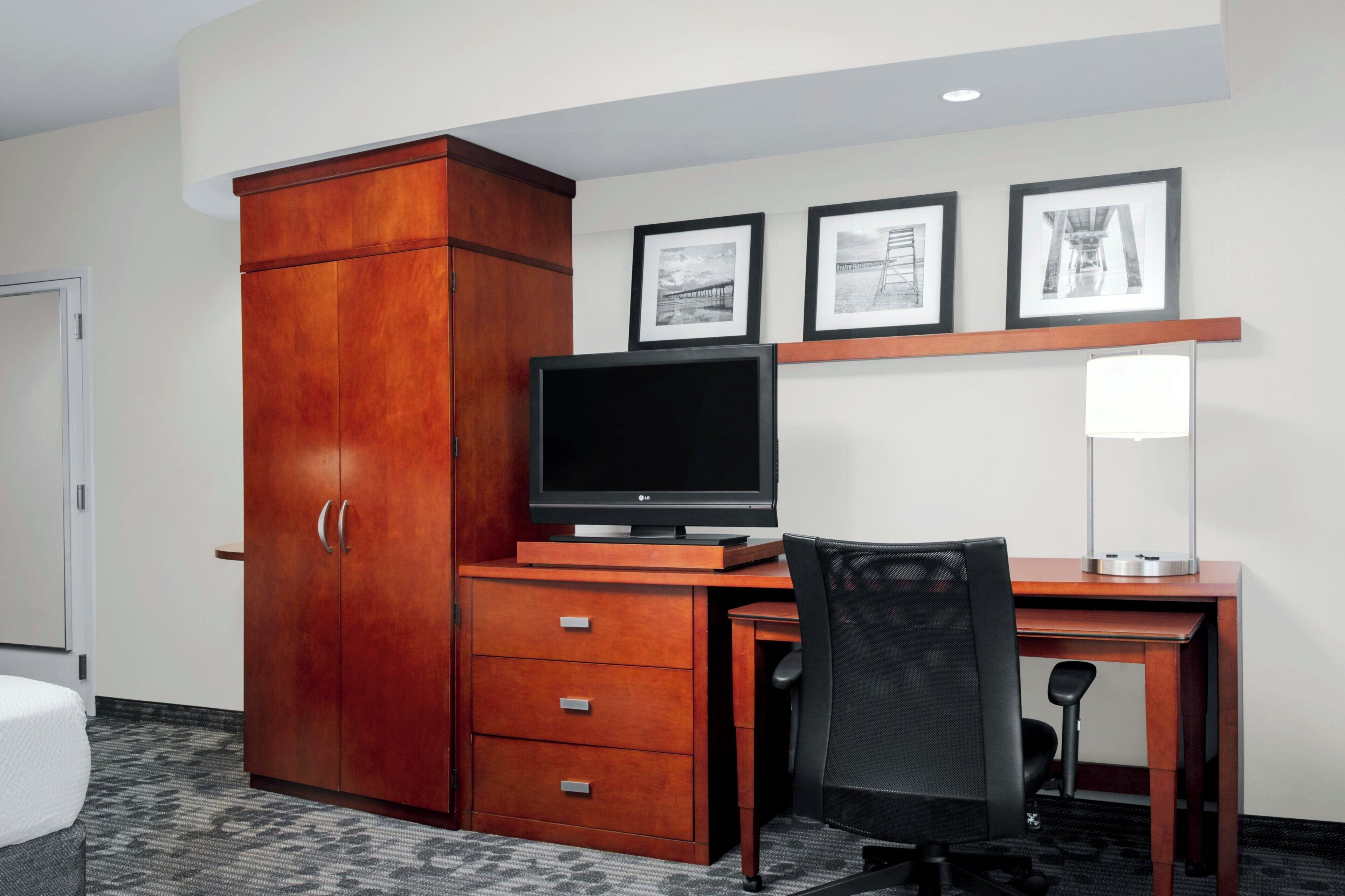 A hotel room with a wooden desk, a computer, a chair, a wardrobe, and three framed pictures on the wall.