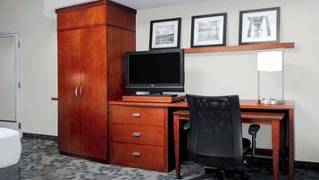 A hotel room with a wooden desk, a computer, a chair, a wardrobe, and three framed pictures on the wall.
