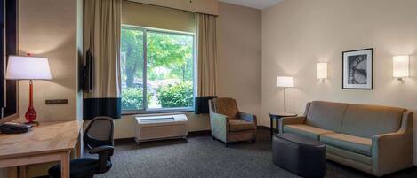 A hotel room with a desk, office chair, sofa, armchair, and a large window with curtains.
