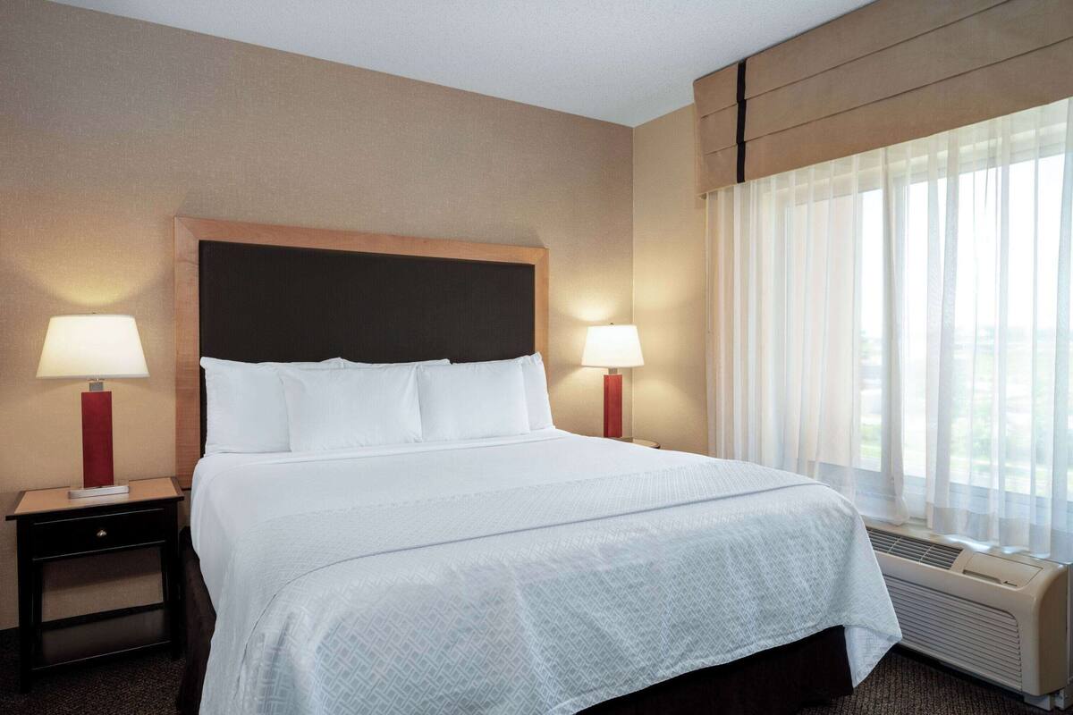 Une chambre d’hôtel avec un grand lit, deux tables de chevet avec des lampes, une fenêtre avec des rideaux légers et un climatiseur fixé au mur.