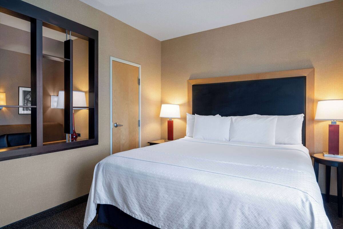 Une chambre d’hôtel avec un grand lit, une tête de lit en bois, deux tables de chevet avec des lampes, une porte et un miroir au mur.
