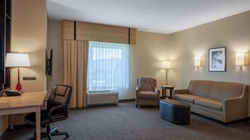 A hotel room with a desk, two chairs, a sofa, a small table, and a wall-mounted clock.