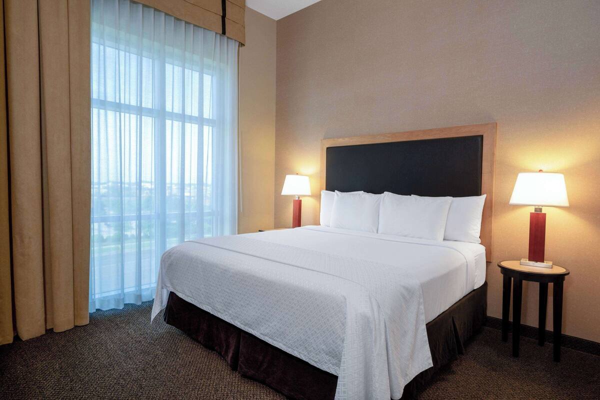 Une chambre d’hôtel avec un grand lit, deux tables de chevet avec des lampes, une fenêtre avec des rideaux légers et une vue sur le paysage urbain.