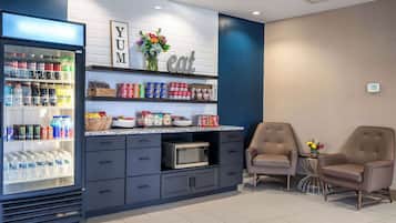 A modern kitchenette with a refrigerator stocked with beverages, a microwave, and various food items on shelves. There are two armchairs and a small table with a vase of flowers.