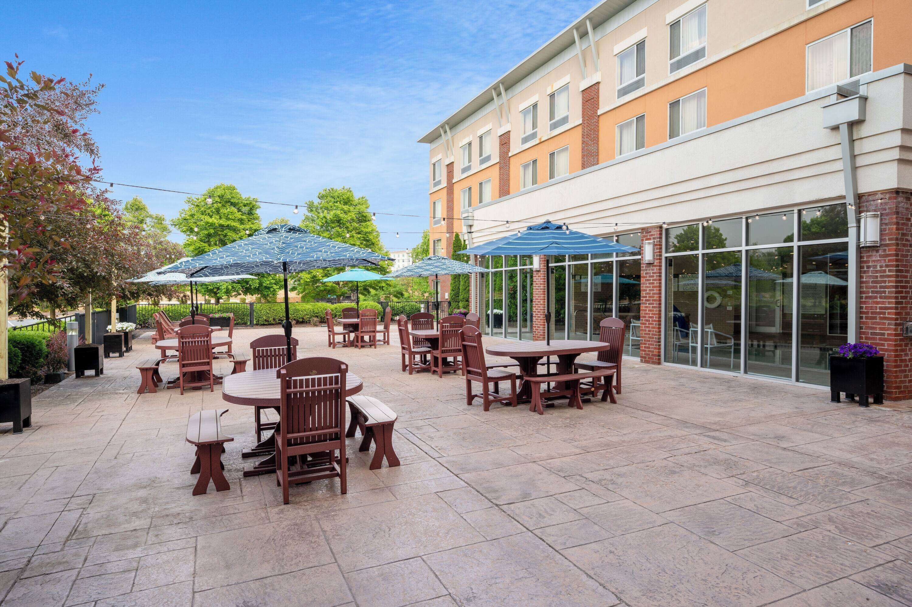An outdoor patio area with tables and chairs, some with umbrellas, and a building with large windows in the background.