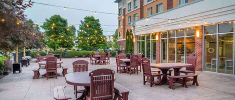 An outdoor patio area with tables and chairs, a building with large windows, and string lights.