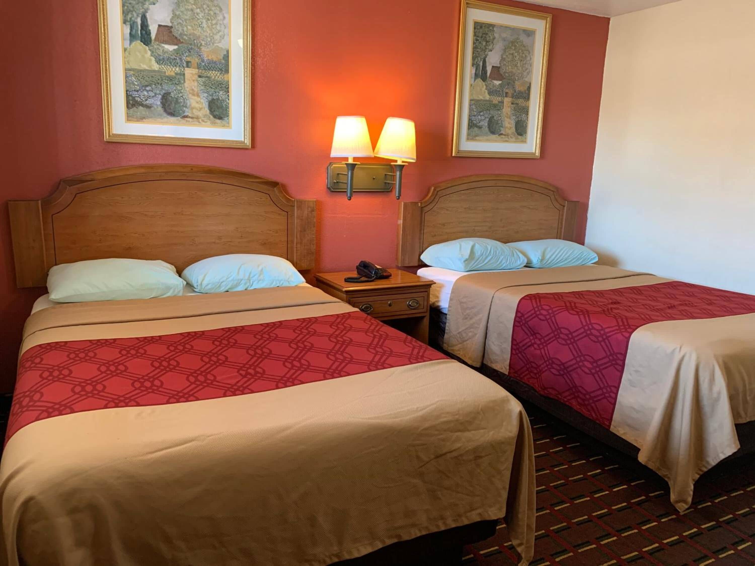 A hotel room with two beds, each with a wooden headboard and a patterned bedspread. There are two wall-mounted lamps on either side of the beds, and two framed paintings above the beds.