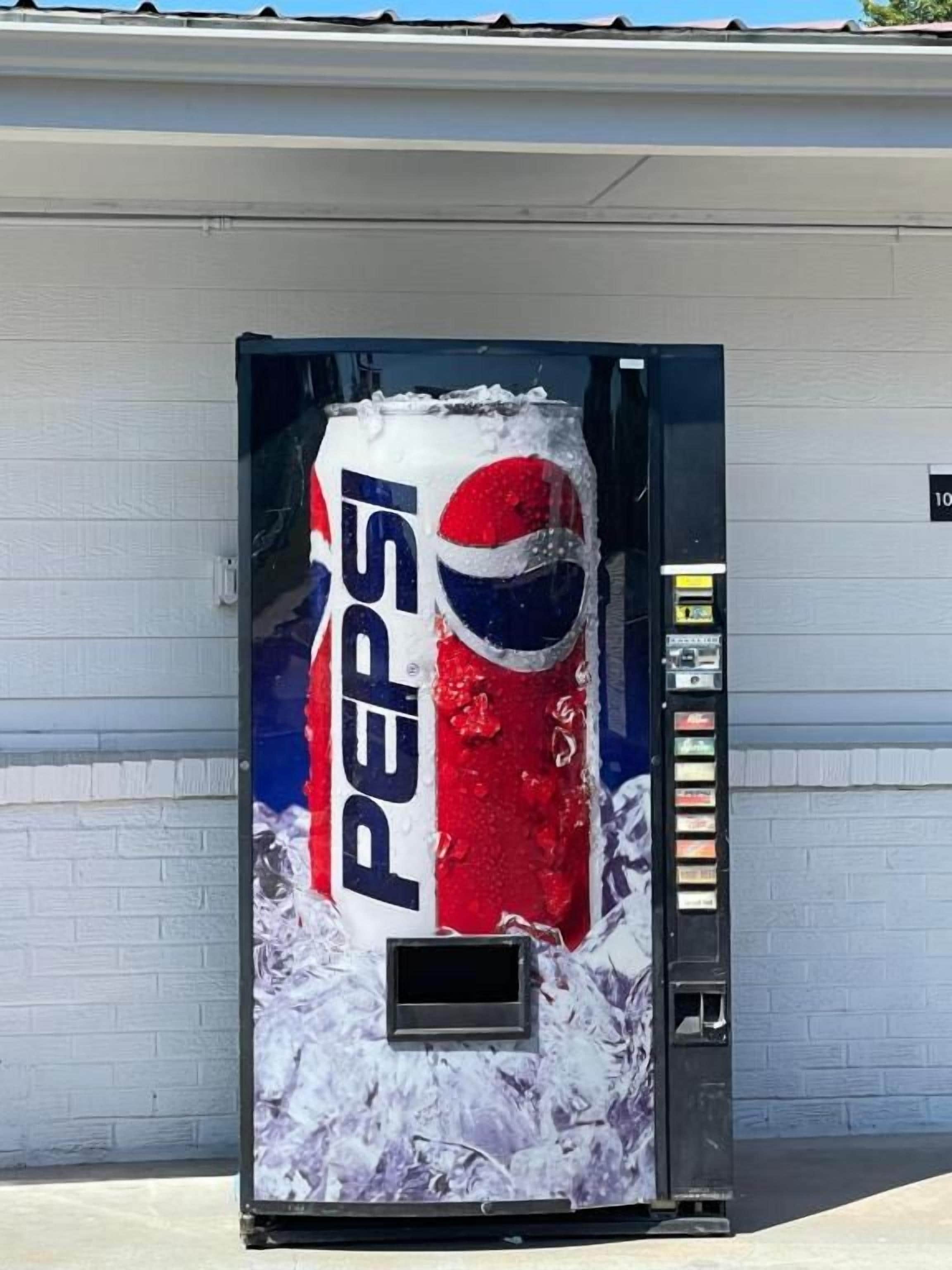 Ein Getränkeautomat mit Pepsi-Logo, vor einem Gebäude mit weißer Wand und schwarzem Eingang.